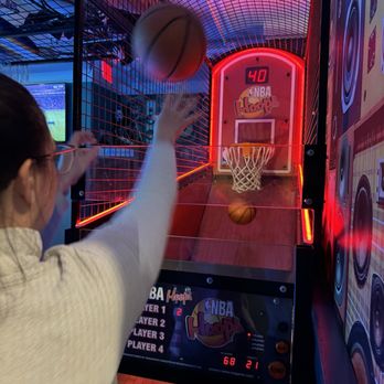 Standard Rec - Gaming Area - A person wearing glasses and a light-colored shirt is playing an arcade basketball game, aiming to shoot a basketball towards the hoop. The game features a digital score display showing 40 points, with neon lighting and a curved ceiling. The venue has a vibrant, colorful atmosphere with a grid-patterned enclosure and wall decorations related to NBA Hoop. The game setup includes a backboard, hoop, and electronic scoring system, indicating a family-friendly entertainment space. - Bars Near Me - Patchogue, New York
