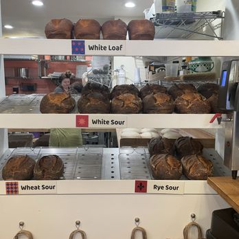 ThoroughBread - Bread Options! - A bakery shelf holds multiple loaves of sourdough bread, each labeled with different varieties such as White Loaf, White Sour, Wheat Sour, and Rye Sour. The loaves are arranged on wire racks, with some appearing freshly baked and crusty. The background shows a bakery kitchen with staff and baking equipment, indicating an active bakery environment. - Bakeries Near Me - Austin, Texas