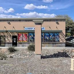 Don't let the outside fool you! - The photo depicts the storefront of AZ Reef Monster, a pet service business in Mesa, Arizona. The building has a beige exterior with large windows displaying colorful posters. The sky above is bright blue with scattered white clouds. The foreground includes a sidewalk and a small landscaped area with rocks and desert plants. The image is taken during daytime with natural lighting.