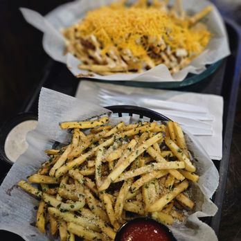 Columbus Axe Throwing - A basket filled with seasoned French fries garnished with herbs and grated cheese, accompanied by a small cup of ketchup and a container of dipping sauce, placed on a dark table with napkins and utensils nearby. - Axe Throwing Near Me - Columbus, Ohio