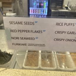 Dry toppings - A menu sign displays Japanese seasoning ingredients such as sesame seeds, rice puffs, and seaweed, with gluten-free labels.
