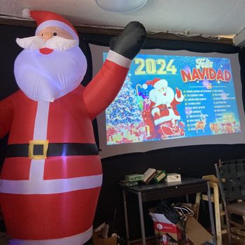 El Comal - a santa claus inflatable in a living room - An inflatable Santa Claus figure stands inside, near a screen displaying a Christmas event schedule for 2024, with festive decorations. - Mexican Near Me - Santa Fe, New Mexico