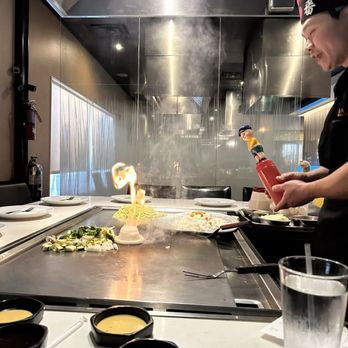 OOKA - Doylestown - A chef is cooking on a teppanyaki grill, with flames and steam visible, in a restaurant setting with plates and condiments nearby. - Japanese Near Me - Doylestown, Pennsylvania
