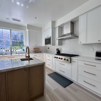 HB Kitchen Bath - The image shows a contemporary kitchen with white cabinets and drawers, a marble backsplash, and a large window allowing natural light. The window has several potted plants on the sill. There is a stainless steel range hood above a gas stove, a microwave built into the cabinetry, and a small countertop appliance. The kitchen island has a marble countertop and a brass faucet. The flooring is light-colored wood, and a dark gray mat is placed in front of the stove. - Kitchen & Bath Near Me - Hayward, California