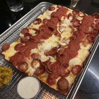 Scarlet Oak - Detroit style pepperoni pizza large - A rectangular Detroit-style pepperoni pizza with a thick crust, topped with melted cheese, pepperoni slices, and tomato sauce, placed on a metal tray. The pizza is accompanied by side condiments, including a small cup of white sauce and a green relish. The setting appears to be a restaurant table with other dishes and drinks visible in the background. - New American Near Me - Washington, DC, District of Columbia