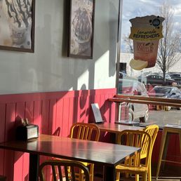 Inside a donut shop, yellow chairs surround a wooden table, with posters on the wall and a window showing parked cars outside.