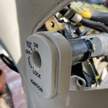 Premier Mobile Locksmith - The image shows a close-up of a motorcycle ignition switch with a key slot, mounted on the motorcycle's frame. The switch has labeled positions: OFF, ON, LOCK, and IGNITION. The surrounding area includes parts of the motorcycle's handlebar and wiring, with some visible dirt and wear. The background is blurred, showing outdoor elements and other vehicles. - Keys & Locksmiths Near Me - Tustin, California