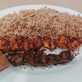 Baba Grill - Lamb and chicken tikka (#19) - The image shows a white oval plate with a serving of seasoned rice, two grilled skewers—one chicken and one beef—and a piece of flatbread on the side. The rice appears fluffy and well-cooked, while the skewers are charred from grilling. The flatbread is rectangular with a slightly toasted surface. The setting suggests a casual dining environment, possibly at a restaurant offering Middle Eastern or South Asian cuisine. - Afghan Near Me - Cinnaminson, New Jersey