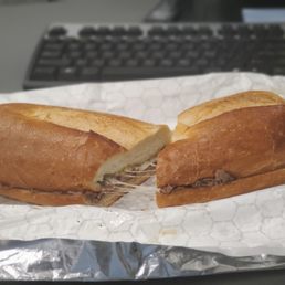 Yorker - A sandwich with sliced roast beef and melted provolone cheese inside a garlic toasted roll. The sandwich is cut in half, showing the filling, and is placed on a piece of wax paper. The background includes a computer keyboard, indicating an indoor setting.