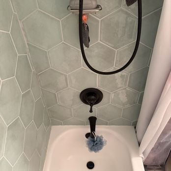 It’s Fixed - I see it every time I get in the shower now - The image shows a shower space with green hexagonal tiles on the walls. A handheld showerhead is mounted on the wall, with a black hose hanging down. A soap dish with a gray soap container is attached to the wall above the showerhead. A gray bath mat with a yellow object is placed on the shower floor, which is white. The shower area is partially enclosed by a white curtain on the right side. - Handyman Near Me