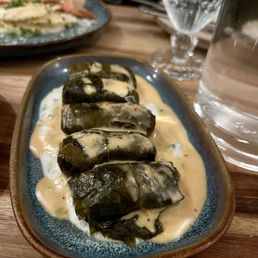 Dolmas - A ceramic plate holds several dolmas covered in a creamy sauce, with a glass of water and a patterned glass in the background.