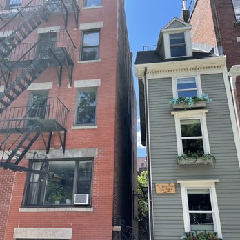 Skinny House - The photo depicts two closely situated narrow buildings in an urban environment. The left building is a red brick structure with a black metal fire escape on its facade. The right building is a light gray, three-story house with window flower boxes filled with greenery. The sky is clear and blue, and the scene captures the architectural contrast between the buildings. - Landmarks & Historical Buildings Near Me - Boston, Massachusetts