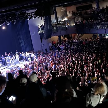 Roadrunner - The suicide file 6/17/2023 View from the standing bleachers - The image shows a spacious indoor music venue filled with a large audience standing and watching a performance on stage. The stage is on the left side, illuminated with bright lights, and has musicians and equipment. The audience extends across the main floor and up to an upper balcony, where additional spectators are present. The venue has a dark interior with visible railings and a sign indicating restrooms. - Performing Arts Near Me - Boston, Massachusetts