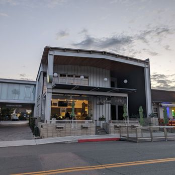 Lofty Coffee - Lofty opens at 0700! - The photo depicts the exterior of Lofty Coffee in Carlsbad, California, with a modern architectural style featuring metal siding and large windows. The open-air seating area is visible with umbrellas and minimal outdoor furniture. The sky is partly cloudy, indicating evening time, and the surrounding area includes a neighboring building with neon signage. The scene is well-lit, emphasizing the contemporary design of the coffee shop. - Coffee & Tea Near Me - Carlsbad, California