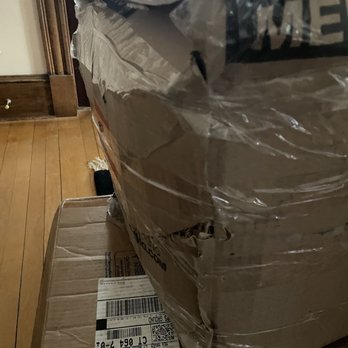 The UPS Store - Damaged boxes deliver to me by UPS - A large cardboard box with torn and crumpled areas is wrapped in clear plastic film. The box is placed on a wooden floor, with a wall and a doorframe visible in the background. The box appears to be damaged, with some parts of the cardboard showing tears and dents, indicating rough handling or shipping issues. - Shipping Centers Near Me - Prospect, Connecticut