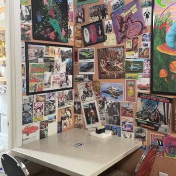 Grumpy's - A small, square table with three chairs is situated in a corner of a room. The walls are densely covered with posters, photographs, and artwork, creating a collage-like display. The ceiling has additional images attached, and a tissue box is on the table. The room appears to be a personal or hobby space with a vibrant, eclectic decor. - American Near Me - Gravette, Arkansas