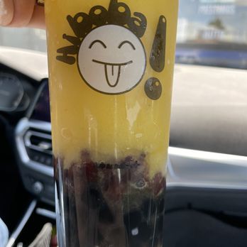 Hi BoBa - A clear plastic cup contains a yellow beverage with dark berries at the bottom. The cup has a sticker featuring a cartoon face with closed eyes, a smiling mouth, and its tongue sticking out, along with the word 'yummy' in a playful font. The drink is being held inside a vehicle, with the dashboard and windshield visible in the background. - Bubble Tea Near Me - Albuquerque, New Mexico