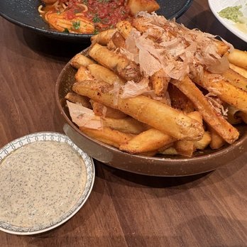 Blu Ember - A bowl filled with French fries seasoned with spices and topped with bonito flakes is placed on a wooden table. A small dish of creamy dipping sauce with herbs is positioned nearby. In the background, other dishes including pasta and salad are partially visible, indicating a dining setting with Asian-inspired cuisine. - New American Near Me - Queens, New York