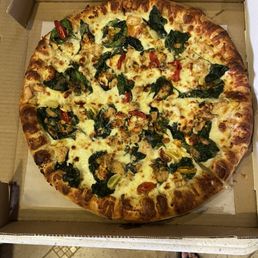 Crazy Californian - A round pizza with a golden-brown crust, topped with melted cheese, fresh spinach leaves, cherry tomato slices, and pieces of cooked chicken. The pizza is placed inside a cardboard box, viewed from above, with a person's feet visible at the bottom of the image.