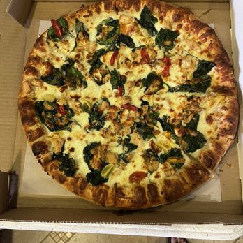 Taboo Pizza - Crazy Californian Pozza - A round pizza with a golden-brown crust, topped with melted cheese, fresh spinach leaves, cherry tomato slices, and pieces of cooked chicken. The pizza is placed inside a cardboard box, viewed from above, with a person's feet visible at the bottom of the image. - Pizza Near Me - Ogden, Utah
