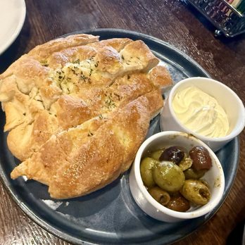 Entourage - A metal plate contains a serving of garlic bread topped with herbs, accompanied by a small bowl of butter and another bowl of marinated green and black olives. The plate is placed on a dark wooden table, with part of a white dish and some restaurant utensils visible in the background. - American Near Me - Downers Grove, Illinois