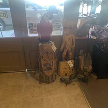 Backroads Art - Two wooden carved figures, one of a colorful parrot and another of a humanoid creature, are placed near the store entrance, with a window and parked cars visible outside. - Art Galleries Near Me - Weston, Missouri
