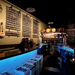 Bar view - A bar interior features a black, curved counter with blue accent lighting underneath. Behind the bar, a large chalkboard menu displays various drink options in white chalk. The wall is decorated with hanging barrels and a guitar mounted on the left side. There are a few patrons seated at the bar, and a bartender is visible in the background. The overall ambiance is dark and cozy, with warm lighting highlighting the menu and decor.