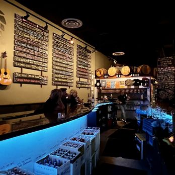 Divided Vine - Bar view - A bar interior features a black, curved counter with blue accent lighting underneath. Behind the bar, a large chalkboard menu displays various drink options in white chalk. The wall is decorated with hanging barrels and a guitar mounted on the left side. There are a few patrons seated at the bar, and a bartender is visible in the background. The overall ambiance is dark and cozy, with warm lighting highlighting the menu and decor. - Wine Bars Near Me - Gilbert, Arizona