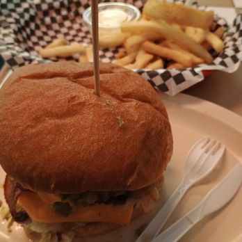 Rex's Hamburgers - Gavster platter - A close-up of a burger featuring a toasted bun, melted cheese, and various toppings, placed on a plate. In the background, there is a basket of French fries with a small container of dipping sauce. The fries are served in a checkered paper-lined basket, and a plastic fork and spoon are on the plate. The setting appears to be a casual dining environment. - Burgers Near Me - Albuquerque, New Mexico