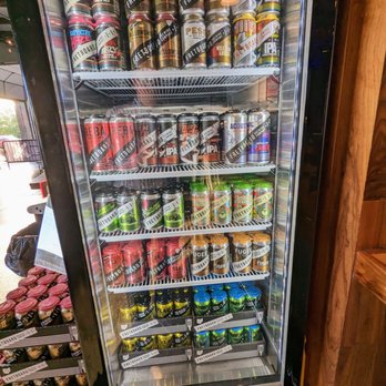 Fretboard Brewing - Beer to Go - A refrigerator with a glass door contains several shelves stocked with cans of craft beer from Fretboard Brewing. The cans are arranged in rows, featuring different flavors and styles, including IPAs, lagers, and specialty brews. The labels display the Fretboard Brewing logo and colorful designs. The fridge is situated indoors, with a wooden wall visible on the right side and a partial view of another display on the left. - Breweries Near Me - Cincinnati, Ohio