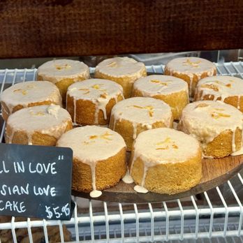Corvus Bakery - 10.01.23 "Fall in Love" Persian love cake - A round wooden tray contains several Persian love cakes with white icing drizzled on top, arranged on a wire shelf in a bakery display. The cakes are small, cylindrical, and have a light golden color. A chalkboard sign in front of the tray reads "Fall in Love" Persian Love Cake, priced at $5.95. The background includes a wooden surface and part of the bakery interior. - Bakeries Near Me - Grass Valley, California