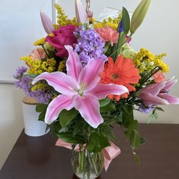 The bouquet - A bouquet of various fresh flowers arranged in a clear glass vase sits on a dark-colored table. The arrangement includes pink lilies, purple stock, orange gerbera daisies, pink roses, and yellow filler flowers, with green foliage. The background features a white wall and a small potted plant. The bouquet is tied with a pink ribbon and appears to be a floral gift or decoration.
