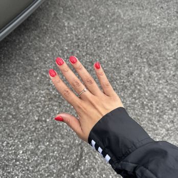 Nail Image - A person's hand with red painted nails and a ring is extended outdoors near a parked vehicle on a textured asphalt surface. - Nail Salons Near Me - Hoover, Alabama