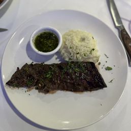 Skirt Steak - A white plate holds grilled skirt steak topped with herbs, mashed potatoes, and a small bowl of chimichurri sauce, set on a white table.