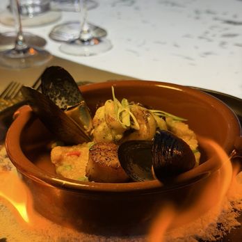Elysium Immersive Dining - Spain - A plated seafood dish includes mussels, potatoes, and microgreens served in a small, round clay pot. The dish is placed on a black tray with a flame underneath, creating a warm presentation. The background shows a white tablecloth and some wine glasses, indicating a fine dining setting. - Modern European Near Me - Lighthouse Point, Florida