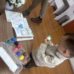 A young boy is engaged in decorating Easter eggs using a paint set with various colors. He is holding a painted egg in his hand and standing at a wooden table that also has a container of paint pots, coloring books, and a flower arrangement. An adult, partially visible, is standing nearby, and the setting appears to be indoors with wooden flooring and white furniture.