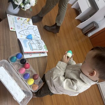 Snap Box - A young boy is engaged in decorating Easter eggs using a paint set with various colors. He is holding a painted egg in his hand and standing at a wooden table that also has a container of paint pots, coloring books, and a flower arrangement. An adult, partially visible, is standing nearby, and the setting appears to be indoors with wooden flooring and white furniture. - Session Photography Near Me - Flushing, New York