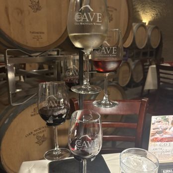 Cave Restaurant - Wine flight - Five wine glasses, containing red and white wines, are stacked in a pyramid formation on a table at Oak Mountain Winery's Cave. The glasses are branded with the winery's logo. In the background, wine barrels are visible, indicating a wine cellar or tasting room setting. A glass of water is also on the table, along with a promotional card for the winery. - Venues & Event Spaces Near Me - Temecula, California