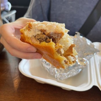 Gomez Salsa - Chicken turtle - A hand holds a partially eaten burrito filled with meat, potatoes, and gravy, with foil wrapping and a takeout container on a wooden table. - Mexican Near Me - Cincinnati, Ohio