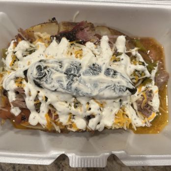 Duke City BBQ - 505 Stuffed Potato - A baked potato is topped with shredded cheese, sour cream, sliced black olives, and possibly other ingredients, served in a white foam container. The toppings are spread over the potato, which appears partially visible beneath the toppings. The container is placed on a surface, likely for takeout or delivery. - Barbeque Near Me - Albuquerque, New Mexico