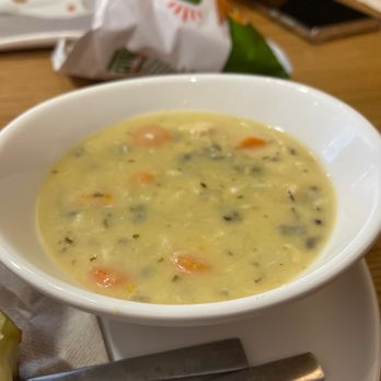Panera Bread - Chicken and wild rice - A bowl of chicken and wild rice soup with visible carrots and herbs is placed on a white plate with a spoon, on a wooden table at Panera Bread. - Sandwiches Near Me - Vestal, New York