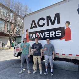 Three men stand side by side in front of an ACM Movers truck on a paved street, each giving a thumbs-up gesture. The truck features a large logo with stylized illustrations of two people and the text 'ACM MOVERS.' The background includes residential buildings with balconies and leafless trees, indicating a possible early spring or late fall setting. The sky is clear and blue.