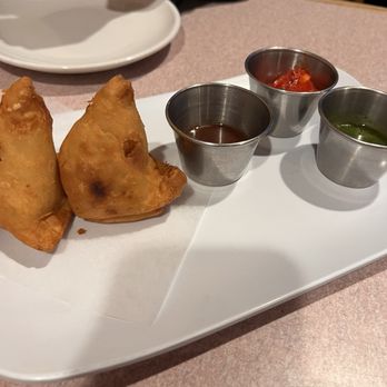 Masala - Vegetarian Samosa - Two vegetarian samosas are presented on a white plate with three small metal cups containing chutneys and sauces, likely at an Indian restaurant. - Pakistani Near Me - West Warwick, Rhode Island
