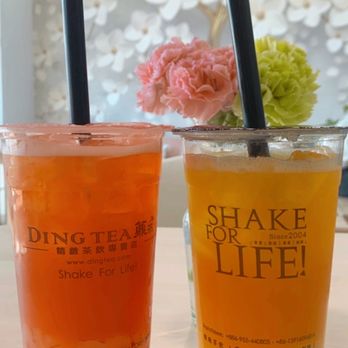 Ding Tea Tucson - Passion fruit green tea - Two fruit teas in clear cups with black straws are placed on a table, with a floral background and a decorative wall. - Bubble Tea Near Me - Tucson, Arizona