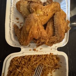 Chicken Wings - The image shows a white, divided takeout container with fried chicken wings in the top section and fried rice with vegetables in the bottom section. The fried chicken wings are golden brown and crispy, while the fried rice appears seasoned and mixed with vegetables. The container is placed on a dark surface, and a fork is partially inserted into the rice.