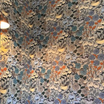 The Corner Counter - Wallpaper is lovely. My favorite so far. - The image shows a wallpaper with a detailed pattern of birds perched on branches among flowers and leaves. The design uses a color palette of blue, gray, black, and orange, creating a nature-inspired motif. The pattern is densely packed, covering the entire visible surface, and the wallpaper appears to be in a well-lit interior space, with a light fixture visible in the upper left corner. - Sandwiches Near Me - Red Hook, New York