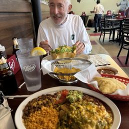 A man with a white beard is eating at a Mexican restaurant, with a plate of rice, beans, and enchiladas, along with a salad and chips.