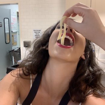 Sur la Table - Me eating pasta - A woman with dark, curly hair and red lipstick is in a restaurant, tilting her head back while holding a fork with long pasta above her open mouth. The background shows white tiled walls, a door, and some signs, indicating an indoor dining environment. - Cooking Classes Near Me - Austin, Texas