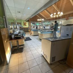 The image shows a buffet restaurant interior with multiple food stations and a person serving food. The ceiling has chandeliers, and the area features tiled flooring and wooden accents. The photo is taken from a vantage point that includes part of the restaurant's interior, with a partial view of the buffet setup and a person in casual clothing.