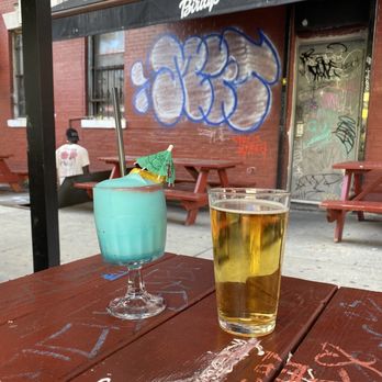 Birdy's - Bud Light and Frozen Margarita - The image shows a blue cocktail with a small umbrella and a straw, alongside a glass of beer, on a wooden table covered with chalk and graffiti markings. In the background, there are red brick walls, graffiti art, and outdoor seating with benches. A person wearing a white shirt and black cap is sitting on a bench, facing away from the camera. The setting appears to be an outdoor area of a bar or pub with an urban, graffiti-covered aesthetic. - Bars Near Me - Bushwick, New York